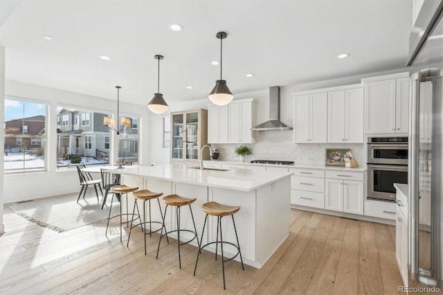 kitchen with appliances with stainless steel finishes, a kitchen island with sink, wall chimney range hood, and white cabinets