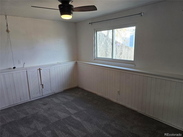 unfurnished room with a ceiling fan, a wainscoted wall, and dark carpet