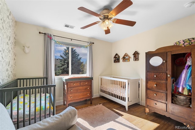 bedroom with a crib, ceiling fan, and dark wood-type flooring