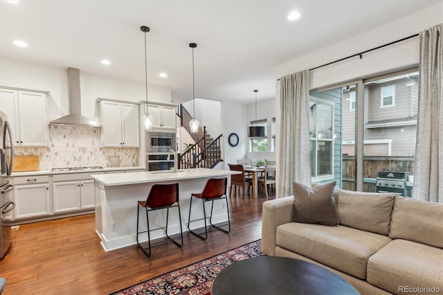 kitchen with wall chimney range hood, appliances with stainless steel finishes, open floor plan, wood finished floors, and recessed lighting