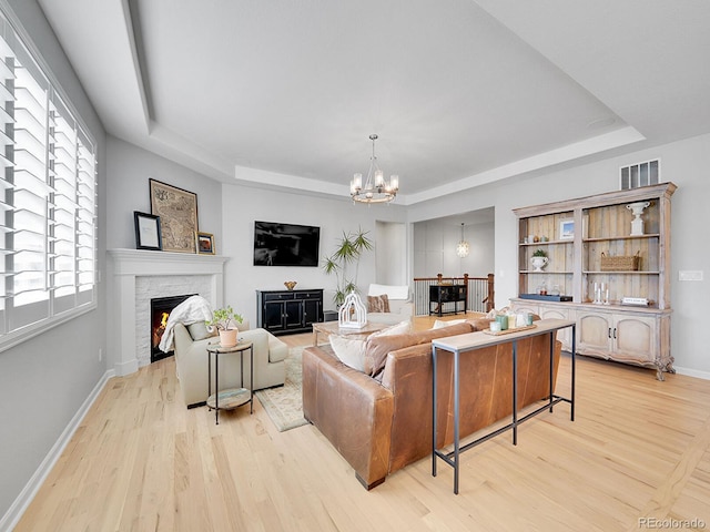 living room featuring visible vents, a stone fireplace, light wood-style floors, a raised ceiling, and a notable chandelier