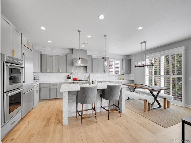 kitchen with light wood finished floors, under cabinet range hood, light countertops, gray cabinets, and a kitchen island with sink