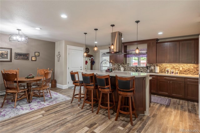 kitchen with island range hood, wood finished floors, a kitchen island with sink, light countertops, and backsplash