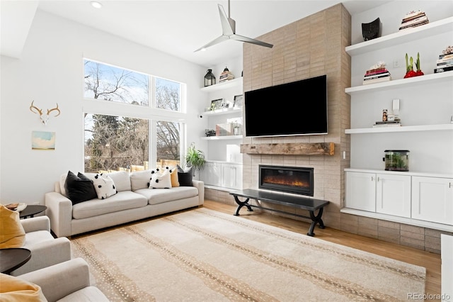 living room with a tiled fireplace, built in shelves, light hardwood / wood-style floors, and ceiling fan