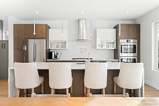 kitchen featuring wall chimney range hood, white cabinetry, appliances with stainless steel finishes, and a breakfast bar area