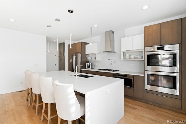 kitchen with an island with sink, white cabinets, hanging light fixtures, stainless steel appliances, and wall chimney range hood