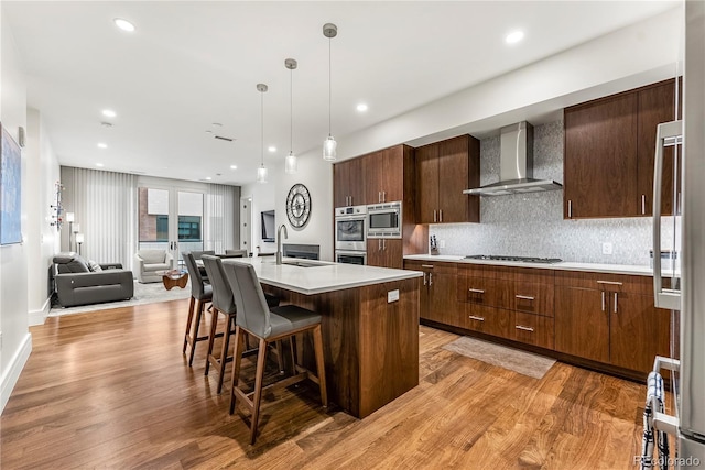 kitchen featuring a breakfast bar, decorative light fixtures, wall chimney range hood, stainless steel appliances, and a center island with sink