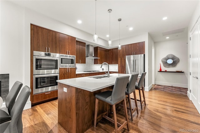 kitchen featuring appliances with stainless steel finishes, pendant lighting, an island with sink, sink, and wall chimney range hood