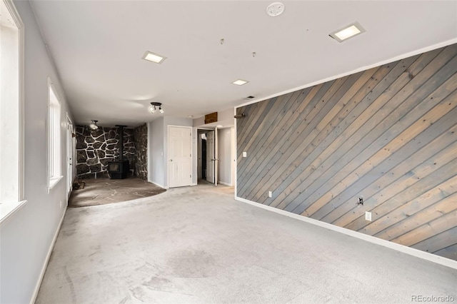 unfurnished living room featuring a wood stove, wooden walls, baseboards, and carpet floors