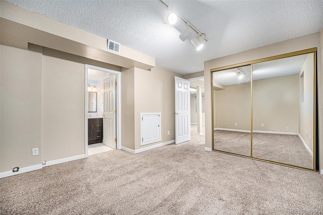 unfurnished bedroom featuring rail lighting, ensuite bathroom, a textured ceiling, light carpet, and a closet