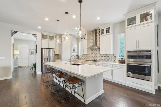 kitchen featuring appliances with stainless steel finishes, white cabinetry, an island with sink, sink, and wall chimney range hood