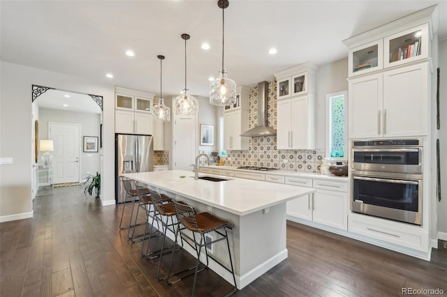 kitchen featuring wall chimney exhaust hood, tasteful backsplash, a kitchen island with sink, a sink, and stainless steel appliances