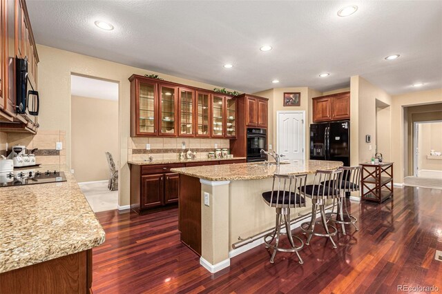 kitchen featuring decorative backsplash, light stone counters, black appliances, a kitchen island with sink, and dark hardwood / wood-style floors