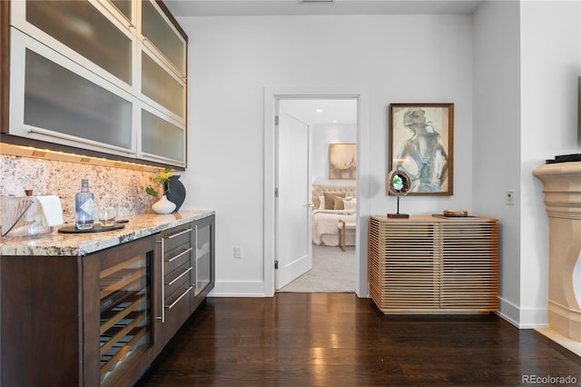 bar with dark wood-type flooring, light stone counters, dark brown cabinets, beverage cooler, and decorative backsplash