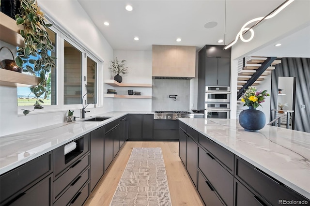 kitchen with appliances with stainless steel finishes, sink, hanging light fixtures, light stone countertops, and light hardwood / wood-style flooring