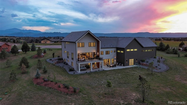 back house at dusk with a balcony, a patio, a mountain view, and a lawn