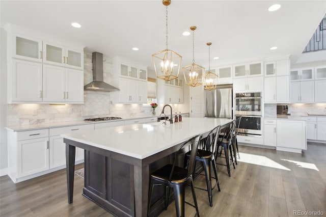 kitchen with wood finished floors, a large island with sink, a sink, appliances with stainless steel finishes, and wall chimney range hood