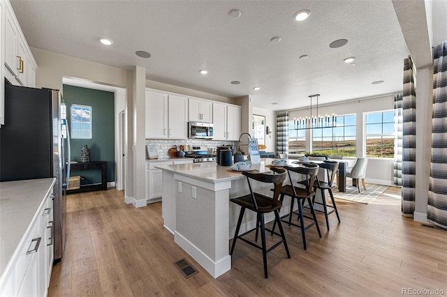 kitchen with tasteful backsplash, an island with sink, white cabinetry, hanging light fixtures, and stainless steel appliances