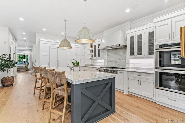 kitchen featuring custom range hood, appliances with stainless steel finishes, white cabinets, light hardwood / wood-style flooring, and a center island with sink