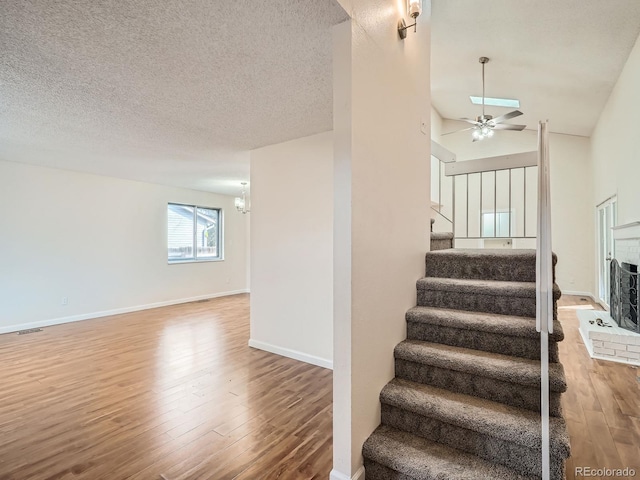 stairway with lofted ceiling, wood-type flooring, a textured ceiling, a brick fireplace, and ceiling fan with notable chandelier