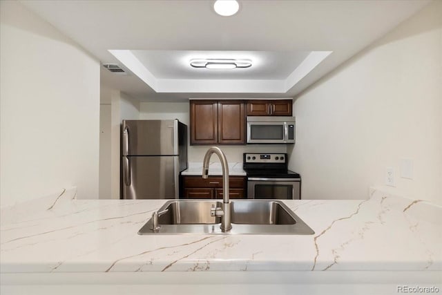 kitchen with stainless steel appliances, a sink, a raised ceiling, light stone countertops, and dark brown cabinets
