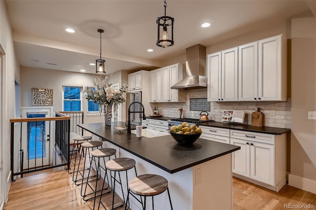 kitchen featuring pendant lighting, wall chimney exhaust hood, light wood-type flooring, and a kitchen island with sink