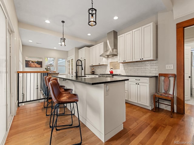 kitchen with hanging light fixtures, wall chimney exhaust hood, an island with sink, and light wood-type flooring