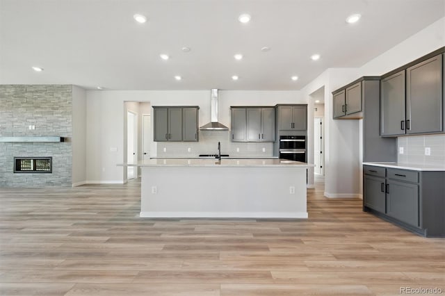 kitchen with a sink, open floor plan, wall chimney range hood, light wood finished floors, and a center island with sink