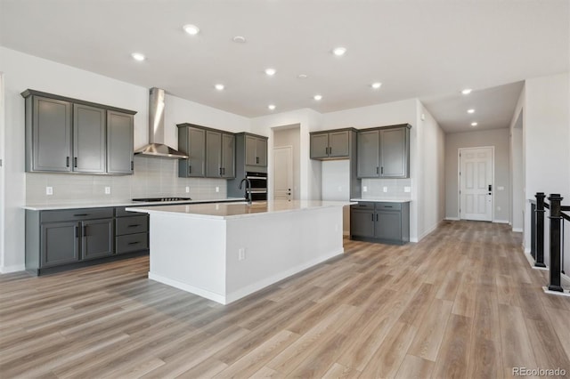 kitchen with gas stovetop, a center island with sink, light wood finished floors, and wall chimney exhaust hood