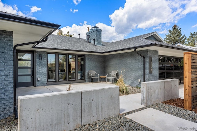 back of property with a chimney, roof with shingles, a patio, and brick siding