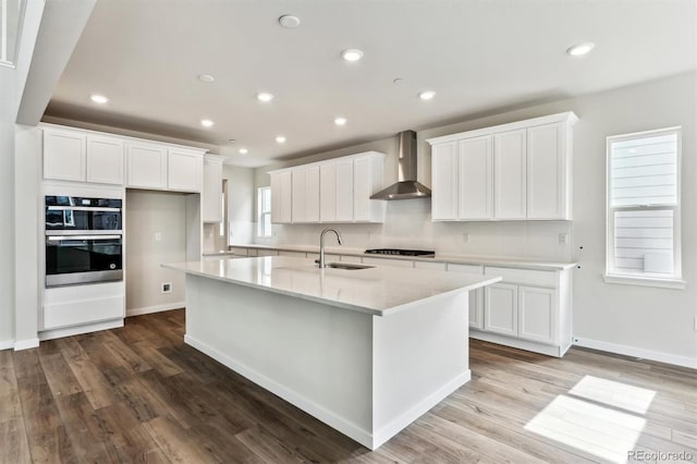 kitchen with an island with sink, a sink, stainless steel double oven, wall chimney range hood, and cooktop