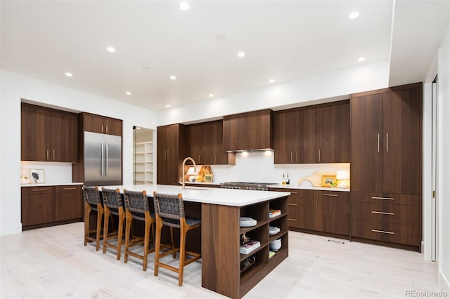 kitchen with built in refrigerator, an island with sink, light hardwood / wood-style floors, a breakfast bar, and dark brown cabinets