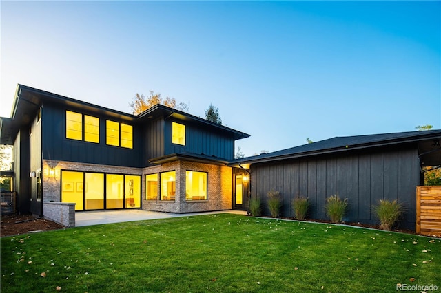 back of house at dusk featuring board and batten siding, a patio area, a lawn, and brick siding