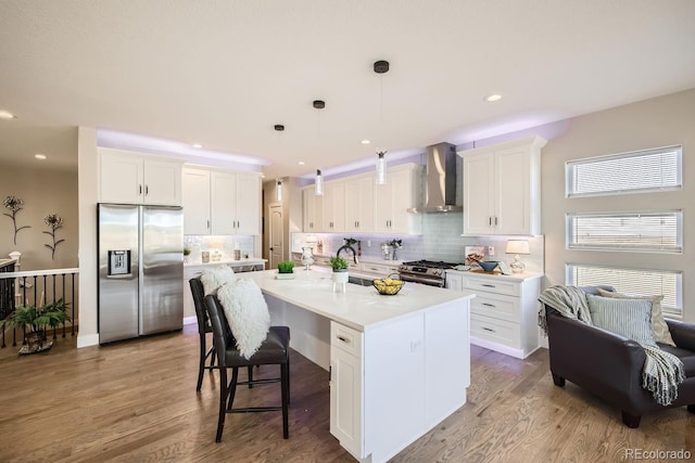 kitchen with white cabinetry, wall chimney range hood, decorative light fixtures, a kitchen island with sink, and appliances with stainless steel finishes