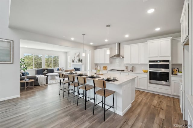 kitchen with wall chimney range hood, stainless steel double oven, light countertops, a kitchen breakfast bar, and open floor plan