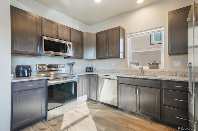 kitchen with dark brown cabinetry, sink, light hardwood / wood-style flooring, stainless steel appliances, and light stone countertops