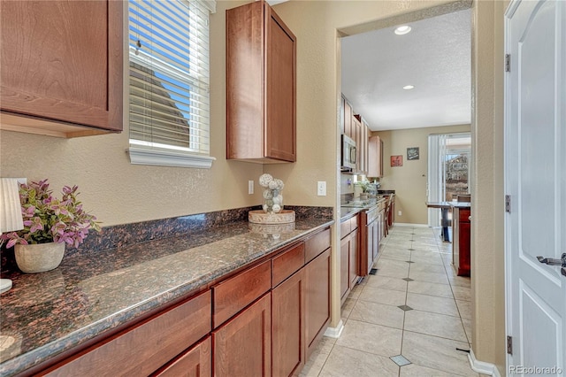 kitchen featuring light tile patterned floors, stainless steel microwave, a healthy amount of sunlight, and dark stone counters