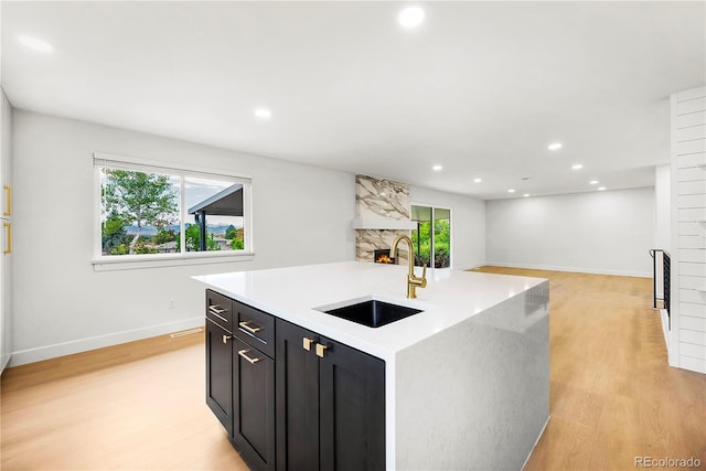 kitchen featuring a high end fireplace, light stone countertops, light hardwood / wood-style floors, a kitchen island with sink, and sink