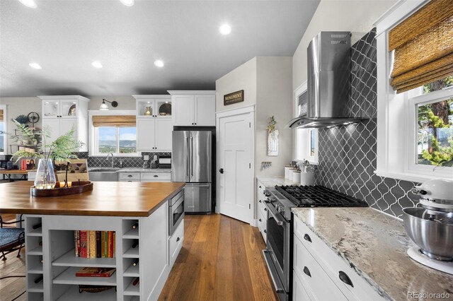 20243 High Pines Dr, Monument, CO 80132 - kitchen with appliances with stainless steel finishes, butcher block counters, wall chimney exhaust hood, backsplash, and white cabinetry