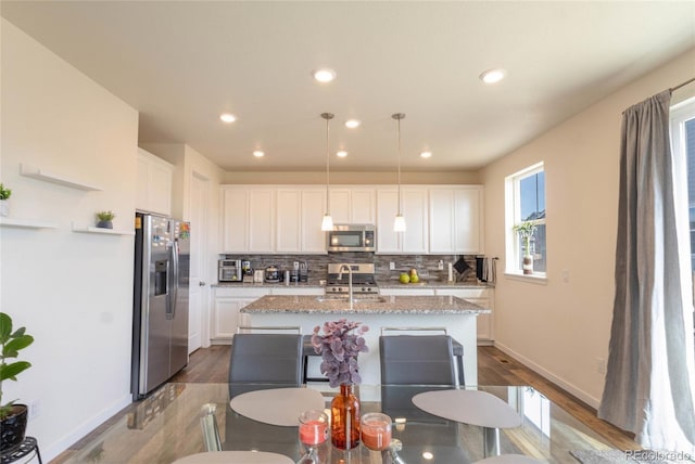 kitchen featuring decorative light fixtures, stainless steel appliances, white cabinetry, an island with sink, and light stone countertops