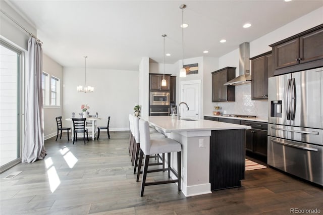 kitchen with appliances with stainless steel finishes, decorative light fixtures, tasteful backsplash, an island with sink, and wall chimney range hood