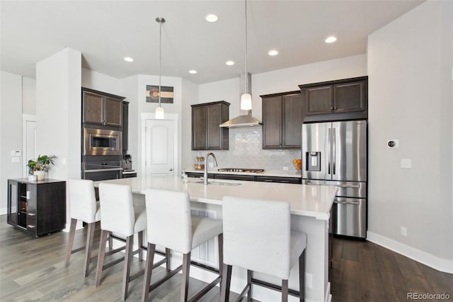 kitchen featuring wall chimney exhaust hood, dark brown cabinetry, decorative light fixtures, an island with sink, and stainless steel appliances