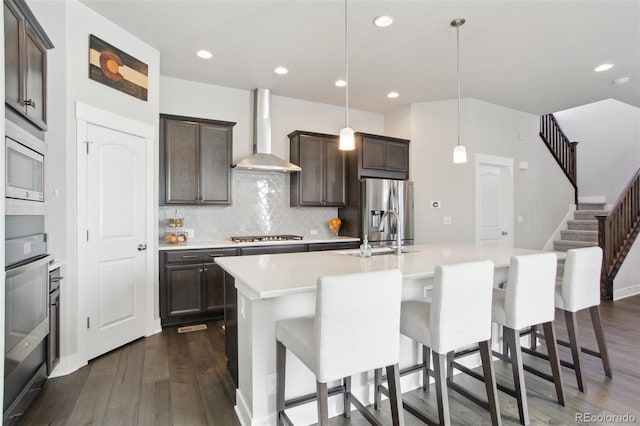 kitchen with wall chimney exhaust hood, sink, hanging light fixtures, an island with sink, and stainless steel appliances