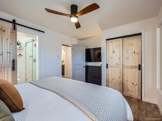 bedroom featuring ceiling fan, a barn door, connected bathroom, dark wood-style flooring, and a closet