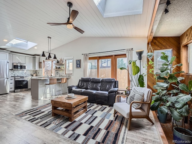 living area featuring vaulted ceiling with skylight, wooden walls, wooden ceiling, ceiling fan, and light wood-type flooring