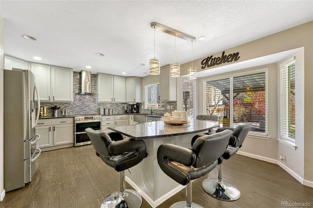 kitchen featuring hanging light fixtures, stainless steel appliances, wood-type flooring, a kitchen bar, and wall chimney exhaust hood