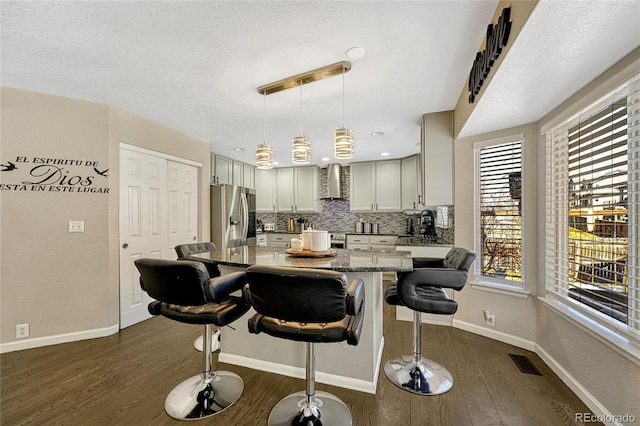 kitchen featuring hanging light fixtures, stainless steel fridge with ice dispenser, light stone countertops, dark wood-type flooring, and wall chimney range hood