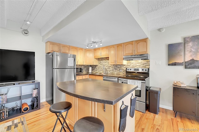 kitchen with dark countertops, tasteful backsplash, under cabinet range hood, light brown cabinetry, and stainless steel appliances