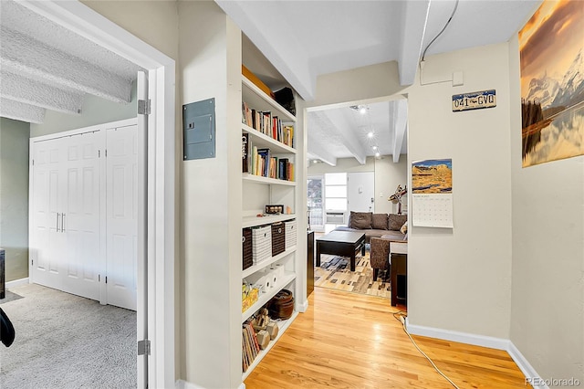 hallway featuring electric panel, beamed ceiling, baseboards, and light wood-style flooring