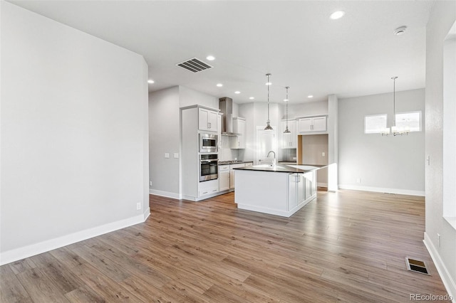 kitchen with stainless steel appliances, wall chimney exhaust hood, a center island with sink, and decorative light fixtures